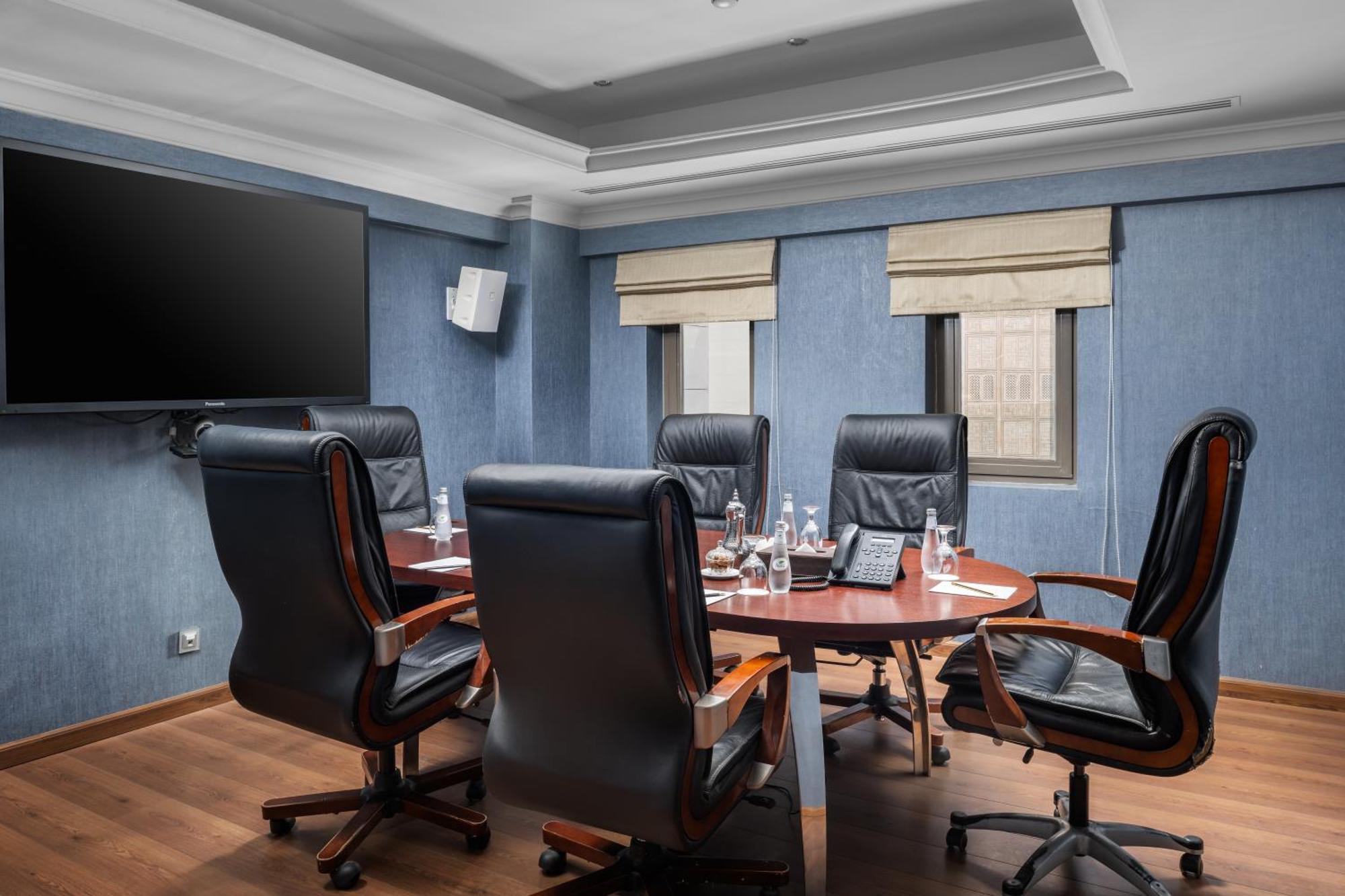 Al Aqeeq Madinah Hotel Medina Exterior foto The photo shows a modern conference room. It features a round wooden table surrounded by several black leather swivel chairs. The walls have a blue color scheme, and there is a large flat-screen TV mounted on one wall. The room has two windows with b