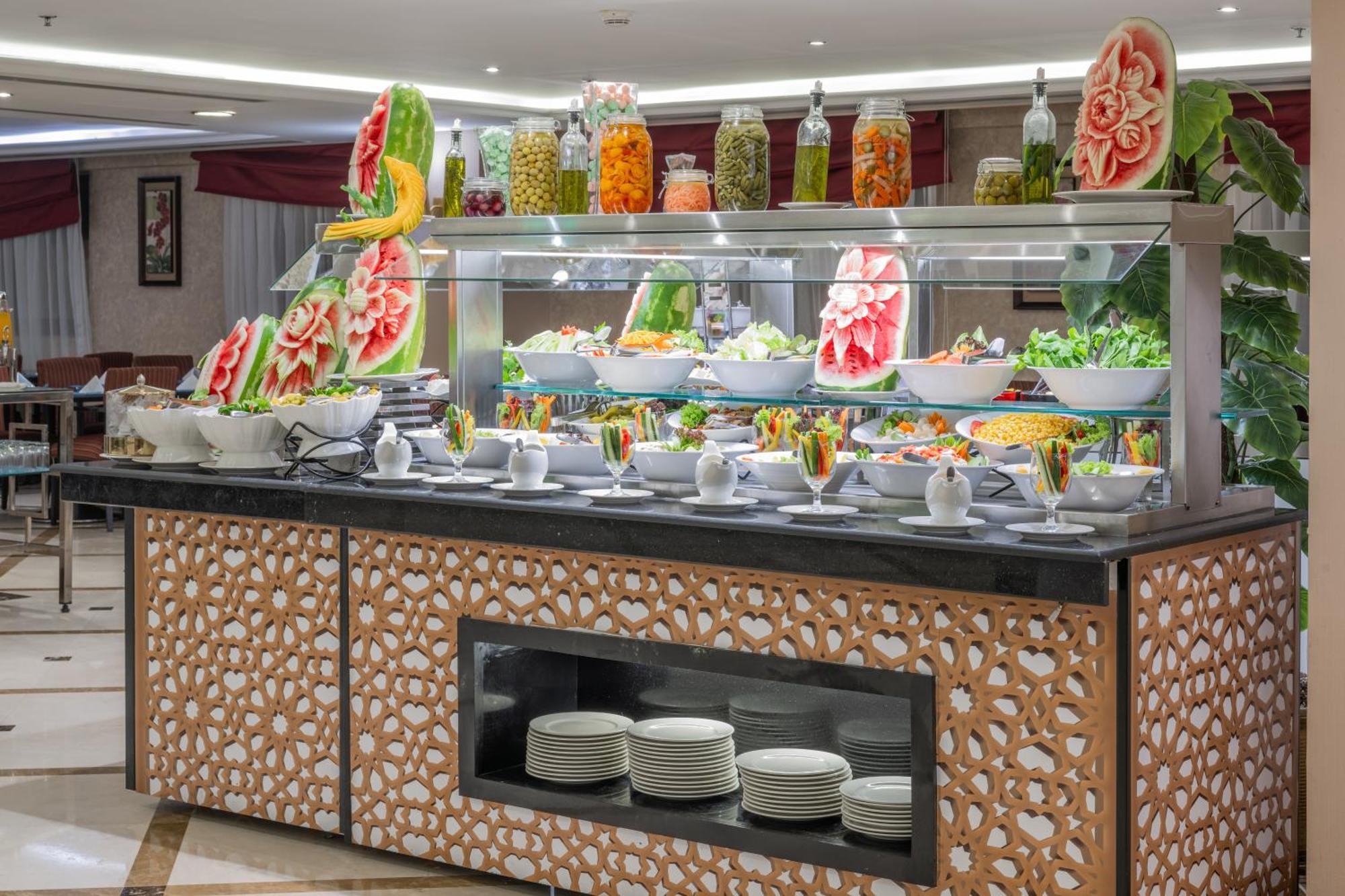 Al Aqeeq Madinah Hotel Medina Exterior foto The photo shows a buffet display that features an array of colorful food items. In the foreground, there are several large bowls filled with various salads and dishes. The presentation is visually appealing, with decorative watermelon carvings and an