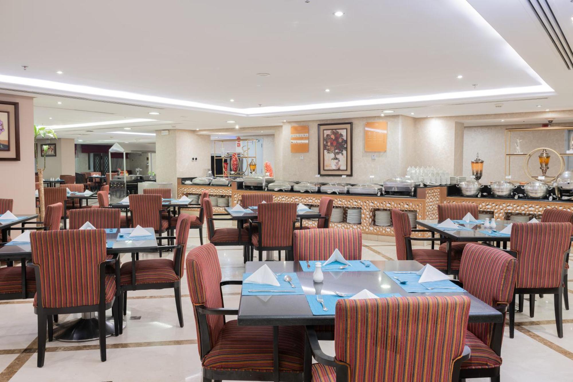 Al Aqeeq Madinah Hotel Medina Exterior foto The photo shows the interior of a restaurant. It features multiple tables arranged neatly, each with patterned chairs and set with blue and white table settings, including folded napkins. The lighting is bright and modern, creating a welcoming atmosp