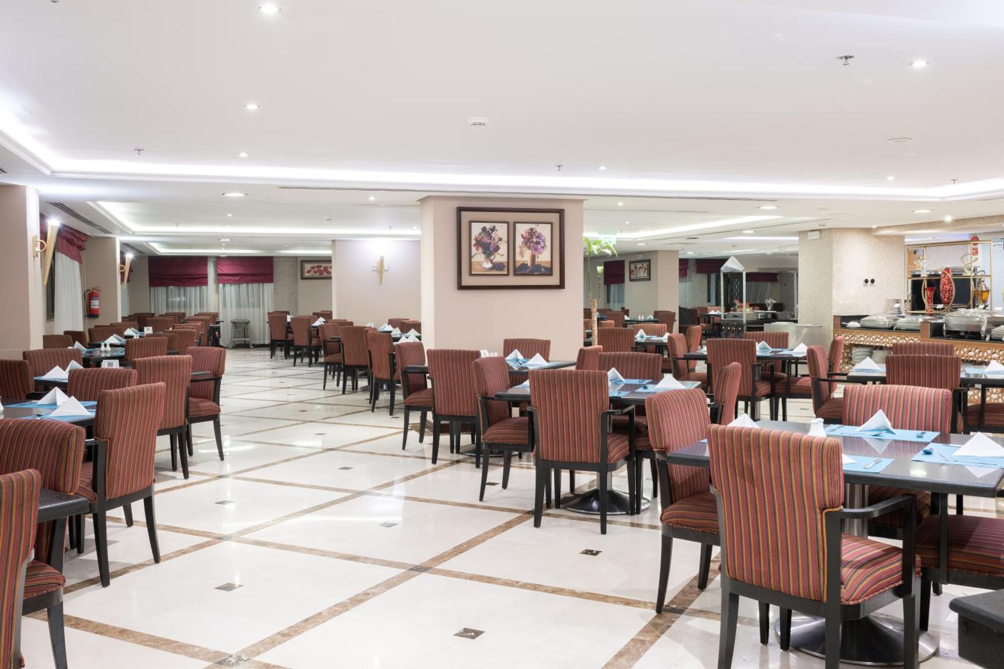 Al Aqeeq Madinah Hotel Medina Exterior foto The photo shows the interior of a spacious restaurant. It features a well-organized dining area with neatly arranged tables and chairs. The tables are dressed with blue napkins, and there is a light-colored flooring with a grid pattern. The walls are