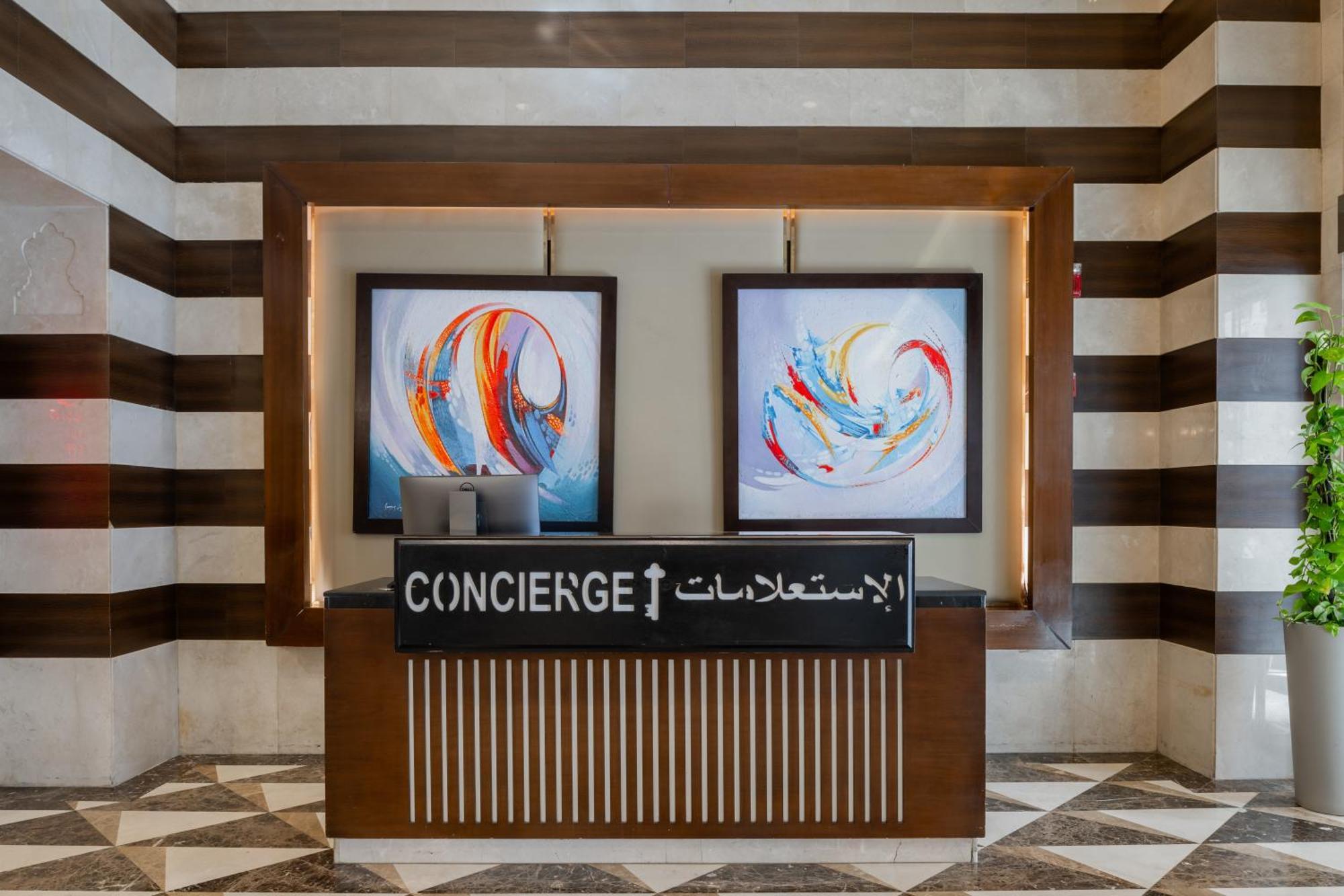Al Aqeeq Madinah Hotel Medina Exterior foto The image shows a concierge desk in a hotel lobby. The desk is made of wood with a striped design and features two large, colorful abstract paintings on the wall behind it. The sign on the desk reads "CONCIERGE" in English and has Arabic text below i