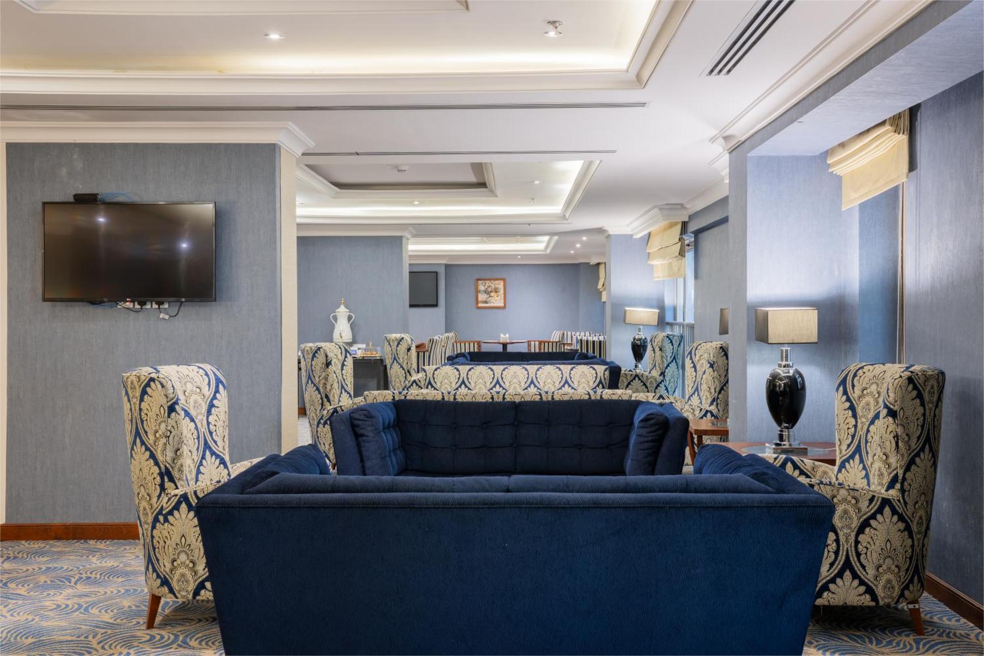 Al Aqeeq Madinah Hotel Medina Exterior foto The photo shows a stylish and modern lounge area. There are two patterned armchairs with blue and cream designs, positioned next to a deep blue couch. The room has a light blue wall with a flat-screen TV mounted on one side. Soft lighting from stylis