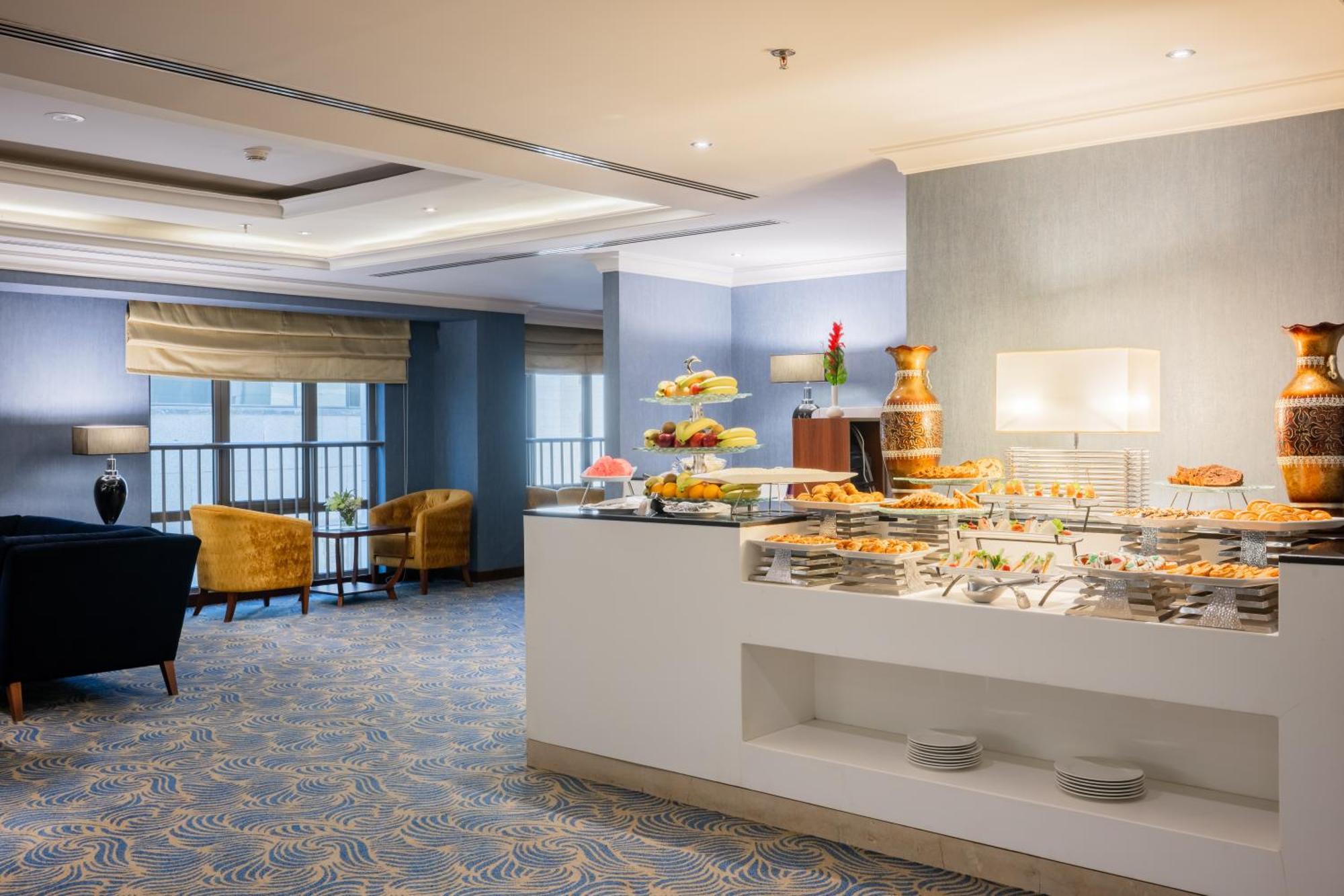 Al Aqeeq Madinah Hotel Medina Exterior foto The photo shows a modern dining area, likely in a hotel or restaurant. On the right side, there is a buffet table with various food items displayed, including pastries and desserts. Behind the table, a decorative fountain is visible. The room feature