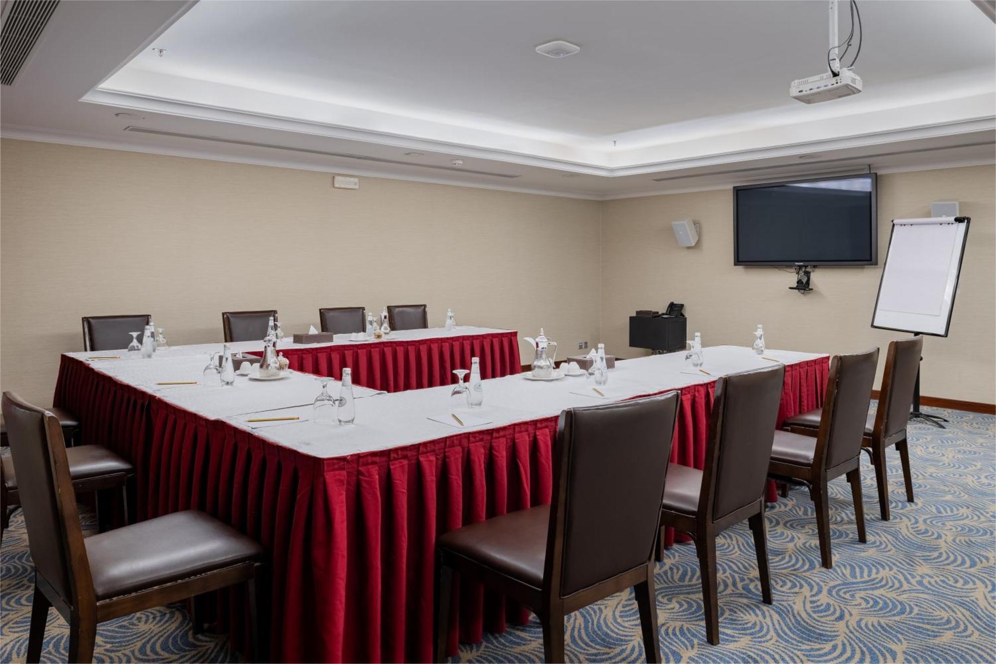 Al Aqeeq Madinah Hotel Medina Exterior foto The image shows a conference room set up for a meeting. There is a U-shaped arrangement of tables covered with white linens and red table skirts. Each table position is equipped with a water bottle and a small notepad, suggesting that it is designed 