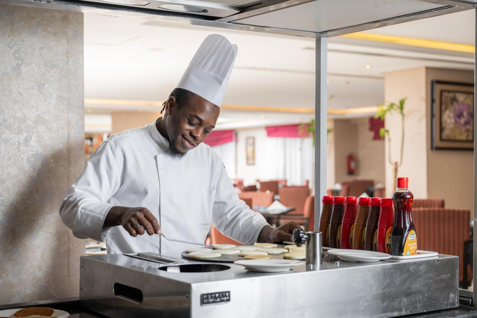 Al Aqeeq Madinah Hotel Medina Exterior foto The photo shows a chef in a professional kitchen setting. He is wearing a traditional white chef's uniform and a tall white chef's hat, and he appears to be smiling while cooking. The chef is standing at a stainless steel cooking station, where he is