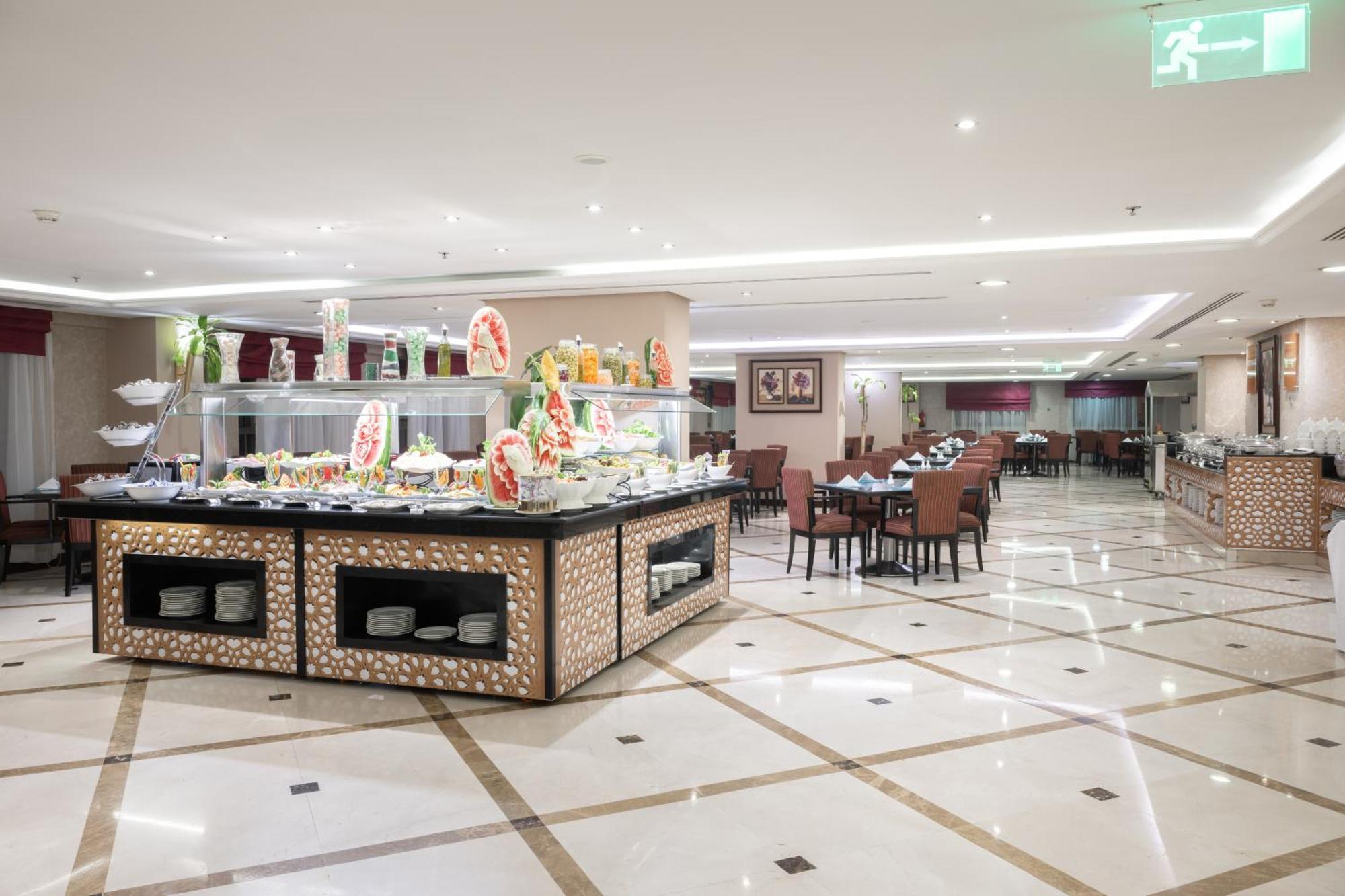 Al Aqeeq Madinah Hotel Medina Exterior foto The photo shows a spacious restaurant interior with a modern design. In the foreground, there is a large buffet counter filled with various food items, beautifully arranged and decorated. The counter features a backlit display, showcasing fruits and 