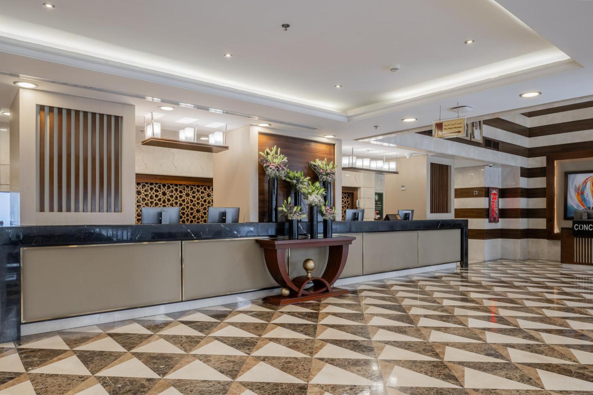 Al Aqeeq Madinah Hotel Medina Exterior foto The photo depicts a hotel reception area. The check-in counter is long and sleek, with a smooth surface and decorative elements. Behind the counter, there are several computer monitors. The walls feature stylish designs, including wooden panels and g