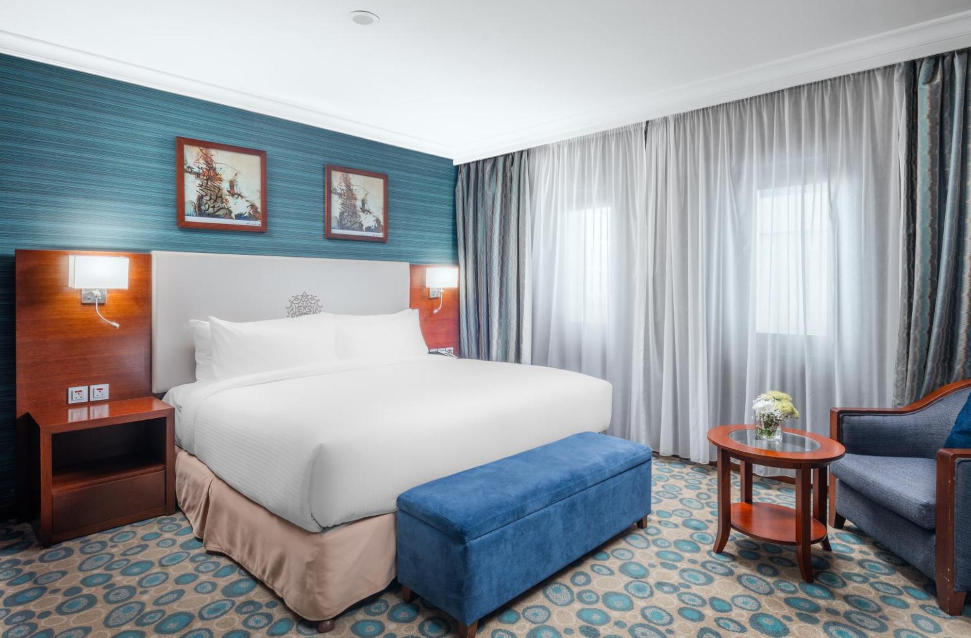 Al Aqeeq Madinah Hotel Medina Exterior foto The photo shows a hotel room featuring a large, neatly made bed with a white duvet. The walls have a light blue accent with framed artwork above the bed. There’s a blue ottoman at the foot of the bed. To one side, there’s a small wooden table with a 