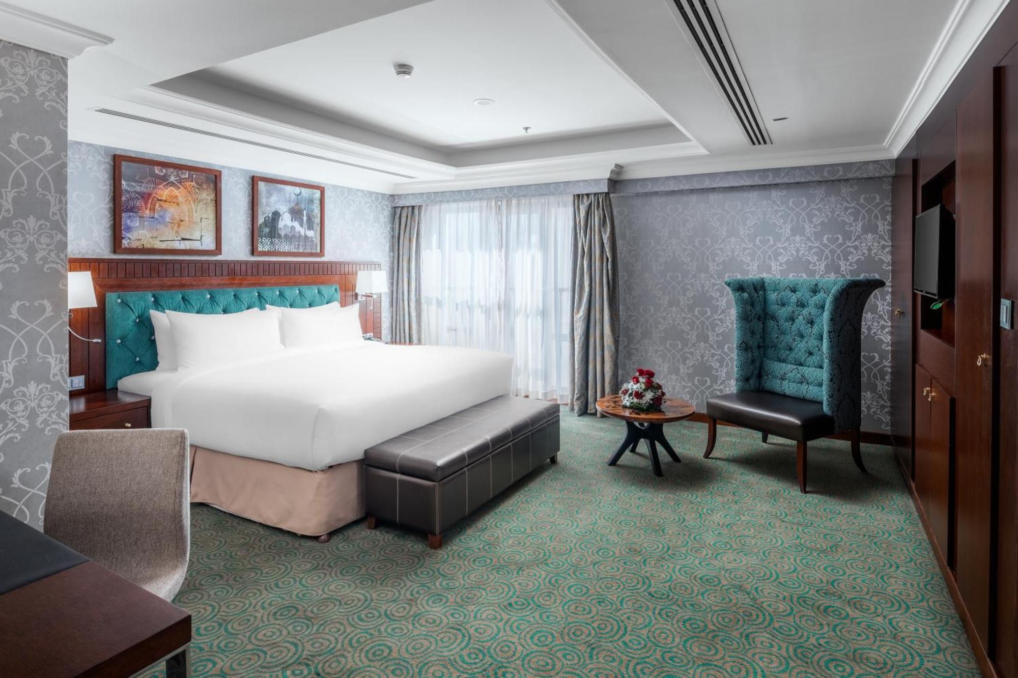 Al Aqeeq Madinah Hotel Medina Exterior foto The photo shows a well-appointed hotel room. It features a large bed with a tufted headboard in a turquoise color, complemented by decorative pillows. There is a comfortable ottoman at the foot of the bed and a small coffee table with a vase of flowe
