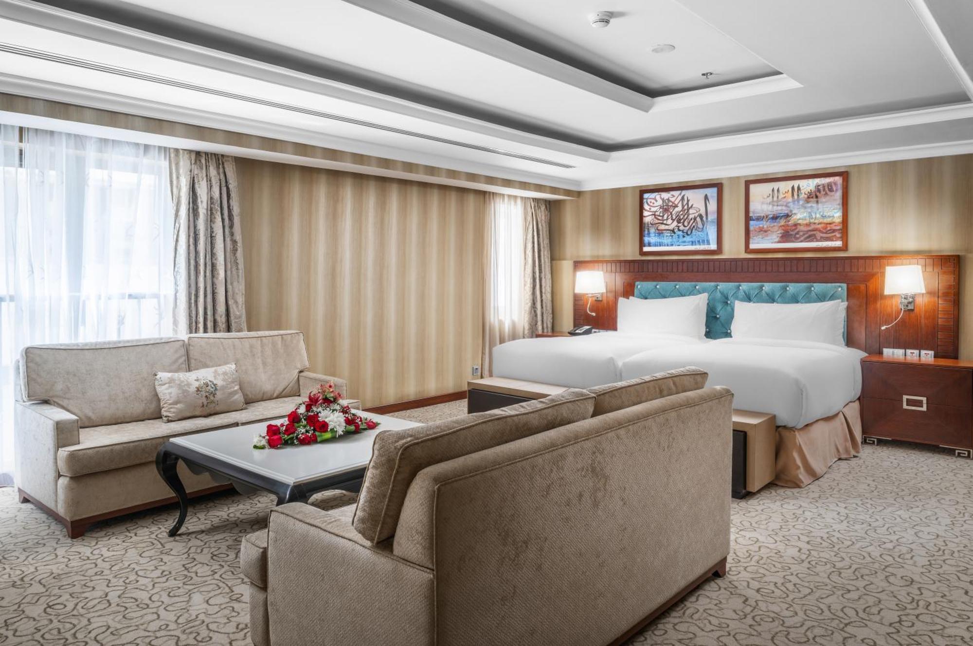 Al Aqeeq Madinah Hotel Medina Exterior foto The photo shows a well-decorated hotel room. It features a large bed with two pillows and a light-colored duvet. There are two comfortable couches arranged around a coffee table with a floral centerpiece. The walls have a subtle striped design, and l