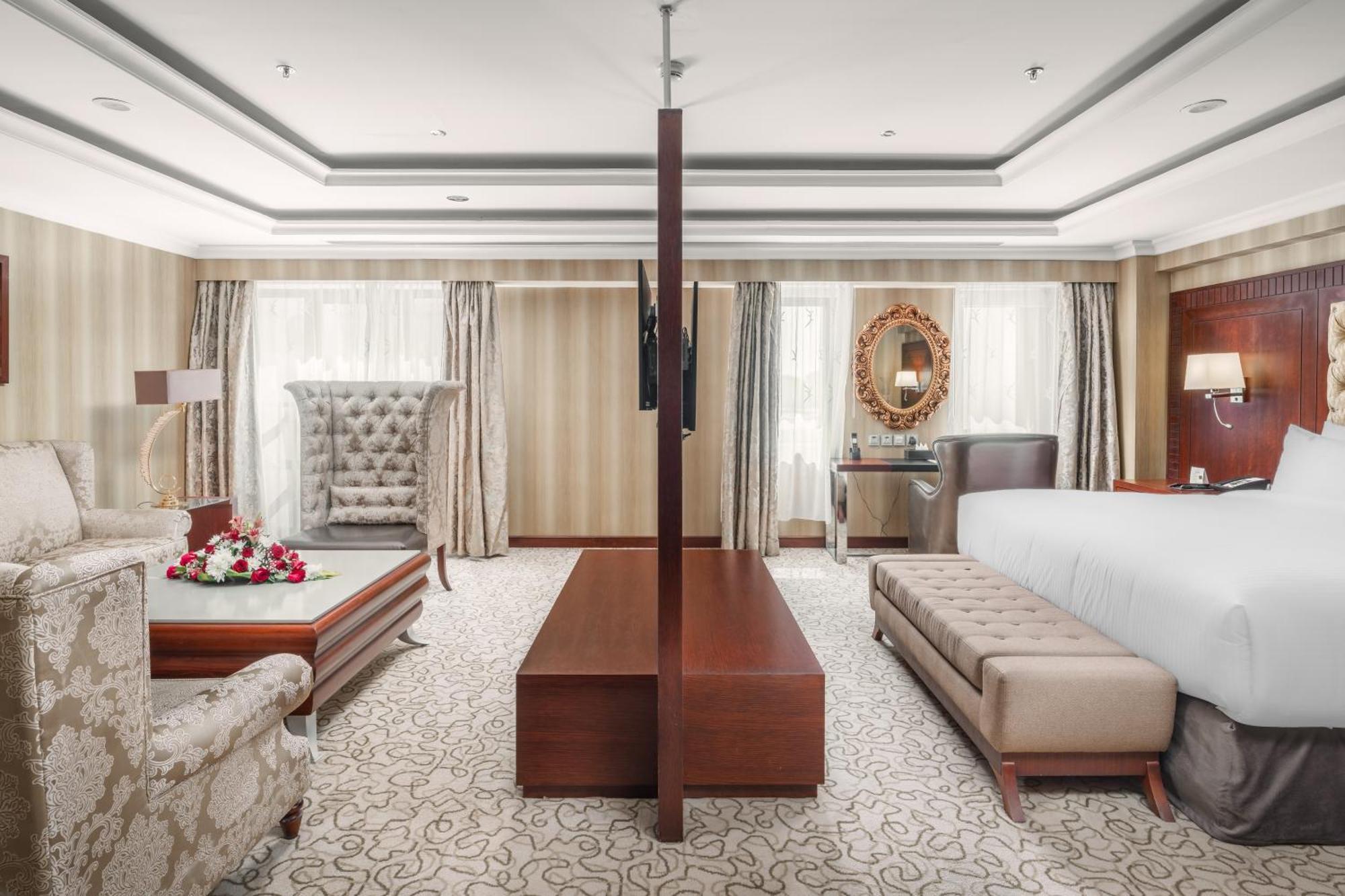 Al Aqeeq Madinah Hotel Medina Exterior foto The photo showcases a spacious hotel suite with an elegant and modern design. The room features a large bed on one side, complemented by a plush bench at the foot. Opposite the bed, there is a stylish seating area that includes a decorative armchair 