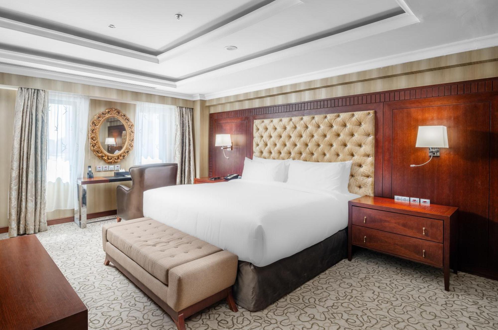Al Aqeeq Madinah Hotel Medina Exterior foto The photo shows a well-furnished hotel room featuring a large bed with a tufted headboard. The room has a warm color palette with wooden accents and soft lighting. There are two windows with curtains that allow natural light to fill the space. Alongs