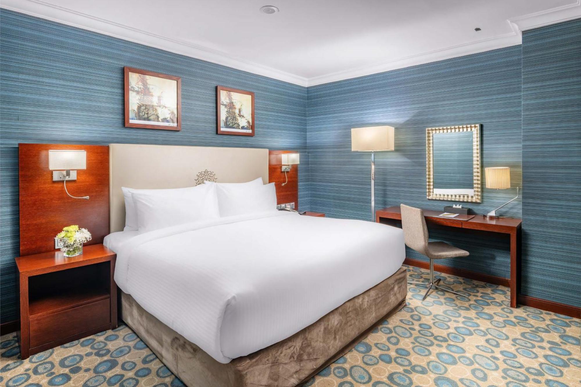 Al Aqeeq Madinah Hotel Medina Exterior foto The photo shows a well-furnished hotel room. It features a large bed with white linens, flanked by wooden nightstands. The walls are painted in a light blue color with a textured pattern. Above the bed, there are two framed pictures. On one side of t