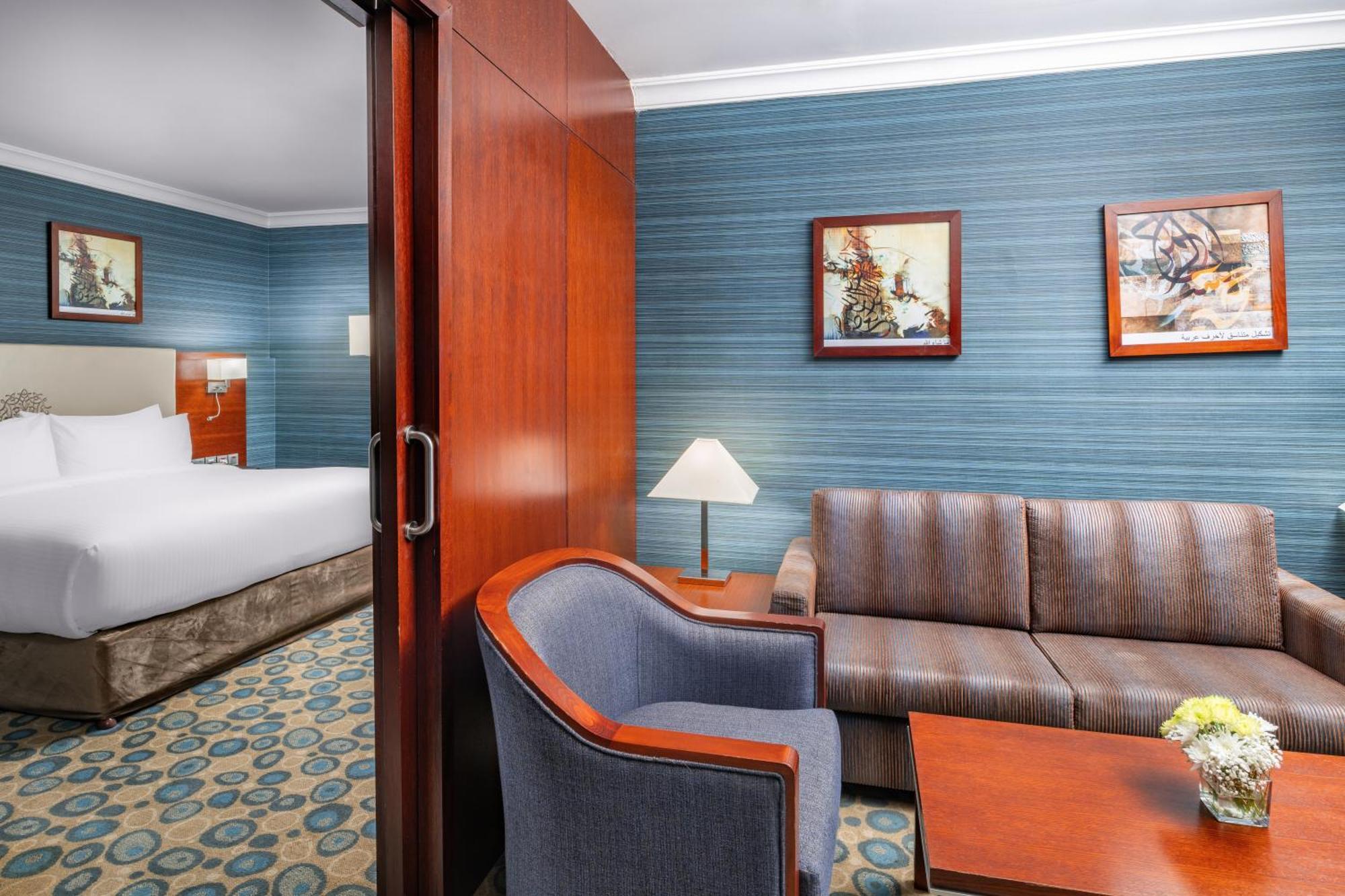 Al Aqeeq Madinah Hotel Medina Exterior foto The photo shows a well-furnished hotel room featuring a living area and a bedroom. To the left, there is a bed visible through an open doorway. The living area includes a gray upholstered chair and a brown couch, both arranged around a small table. A