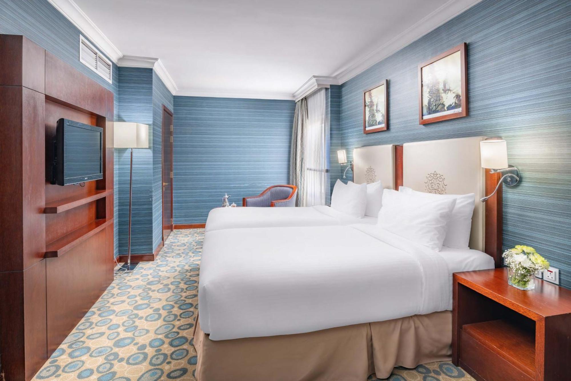 Al Aqeeq Madinah Hotel Medina Exterior foto The photo shows a hotel room featuring two twin beds dressed in white linens. The walls are painted in a soft blue color, and there are framed artworks hung above the beds. A flat-screen TV is mounted on a wooden unit against one wall. To the side, t