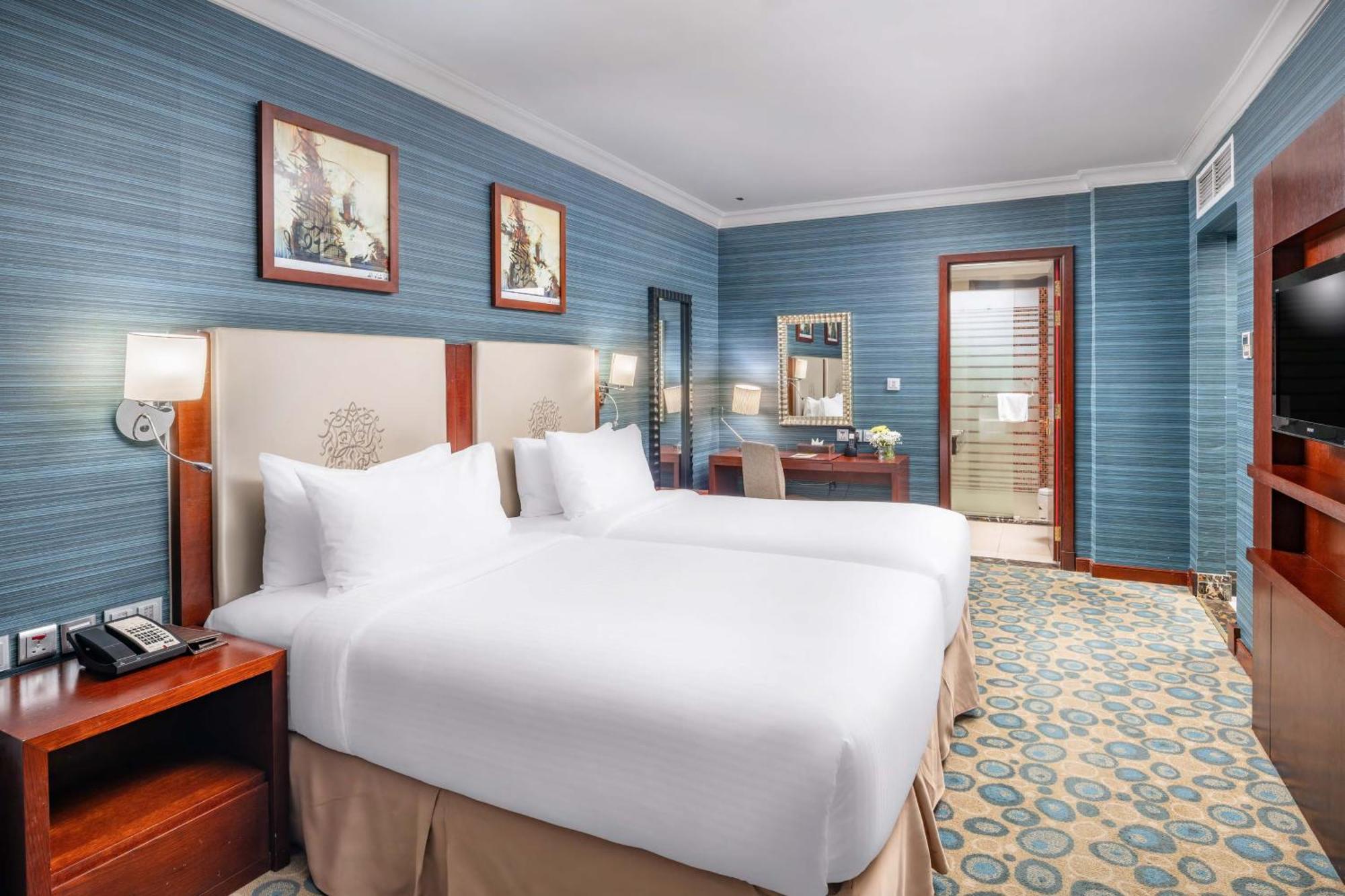 Al Aqeeq Madinah Hotel Medina Exterior foto The photo shows a hotel room featuring two twin beds with white bedding, positioned against a light-colored headboard. The walls are painted in a soft blue shade, adorned with framed artwork. There is a wooden desk with a chair, and a mirror mounted 