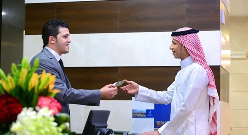 Al Aqeeq Madinah Hotel Medina Exterior foto The image depicts two individuals at a reception desk. The person on the left, dressed in a formal suit, appears to be handing a card or document to the person on the right, who is wearing traditional attire, including a white thobe and a red and whi