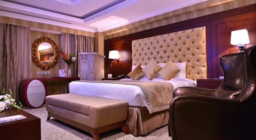 Al Aqeeq Madinah Hotel Medina Exterior foto The photo shows a luxurious hotel room. It features a large, upholstered headboard with a tufted design, paired with plush bedding and decorative cushions. There's a stylish bench at the foot of the bed and a comfortable armchair to the side. The roo