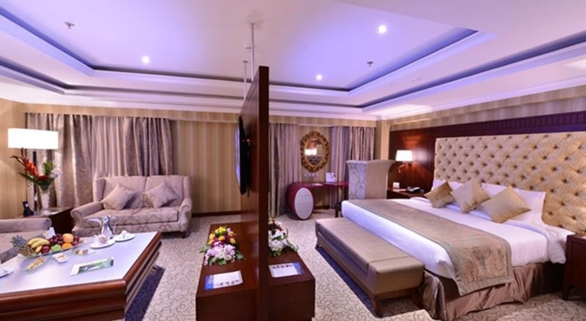 Al Aqeeq Madinah Hotel Medina Exterior foto The image shows a luxurious hotel room. It features a large bed with a tufted headboard, flanked by nightstands with lamps. There's a seating area with a plush sofa and a coffee table. A decorative partition separates the bed from the living area. Th