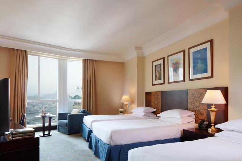 Al Aqeeq Madinah Hotel Medina Exterior foto The photo shows a hotel room featuring three beds arranged neatly. The room has a warm, inviting ambiance with beige walls and a soft carpet. Large windows allow natural light to fill the space, offering a view outside. There are curtains in a light 