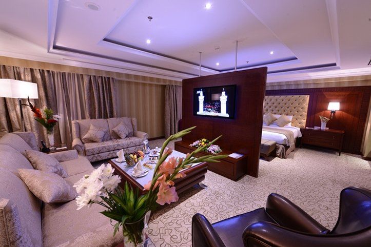 Al Aqeeq Madinah Hotel Medina Exterior foto The photo shows a luxurious hotel room suite. There is a comfortable seating area with stylish sofas and a coffee table. A decorative arrangement of flowers is on the table, adding a touch of elegance. A large flat-screen television is mounted on a p