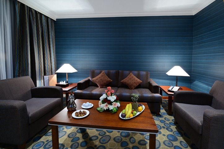 Al Aqeeq Madinah Hotel Medina Exterior foto The photo shows a cozy living area featuring a dark blue wall with a textured design. In the center, there is a wooden coffee table surrounded by two comfortable armchairs and a sofa with decorative cushions. On the table, there are plates with snack