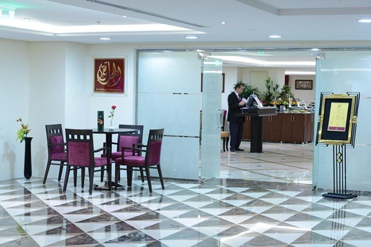 Al Aqeeq Madinah Hotel Medina Exterior foto The photo shows a modern interior space, likely a lobby or reception area of a hotel or restaurant. It features a checkered floor with a combination of dark and light tiles. On the left, there is a small table set for two, adorned with a single rose 