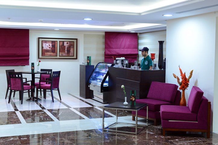 Al Aqeeq Madinah Hotel Medina Exterior foto The photo shows a modern interior space, likely a café or small restaurant. It features a seating area with a few tables and chairs, all in a dark wood finish with purple upholstery. There is a counter area with a staff member behind it, possibly ser