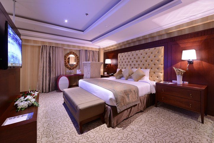 Al Aqeeq Madinah Hotel Medina Exterior foto The photo shows an elegantly designed hotel room. It features a large bed with a plush headboard and decorative pillows. On either side of the bed are bedside tables with lamps. There's a comfortable bench at the foot of the bed and a TV mounted on t