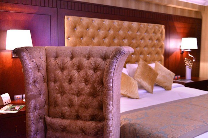 Al Aqeeq Madinah Hotel Medina Exterior foto The image shows a luxurious bedroom featuring a large upholstered headboard with a tufted design in a soft, beige color. The bed is elegantly dressed with decorative pillows and a textured bedspread. In the background, there are wooden paneling and s